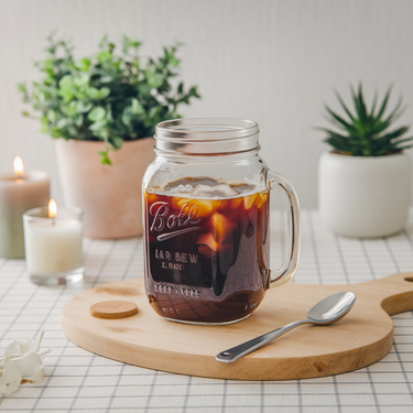 A glass mason jar of iced coffee made from 🦈 Jawesome Cold Brew Pods sits on a wooden board with a spoon, surrounded by candles and potted plants on a checkered tablecloth.